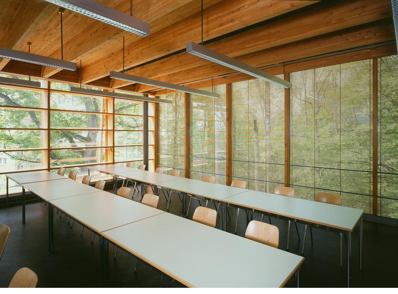 Wiesbaden Fachhochschule classroom interior — glulam timber roof, OKAWOOD glazing on two walls, dappled striped daylight across desks